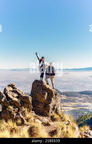 Zwei Freunde stehen auf einem felsigen Gipfel und genießen einen atemberaubenden Blick auf die Berge unter einem klaren blauen Himmel. Ihre fröhlichen Ausdrucksformen fesseln das Wesen von adve Stockfoto
