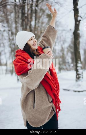Eine fröhliche junge Frau lächelt, als sie in einem verschneiten Park ihre Hand hebt, warm gekleidet mit einem roten Schal, einer weißen Mütze und einer Tasse Kaffee. Stockfoto