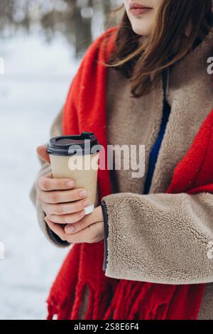 Zugeschnittene Ansicht einer jungen Frau in einem leuchtend roten Schal mit einer Kaffeetasse in einem verschneiten Wald. Ihr Gesicht ist teilweise sichtbar, fokussiert auf ihre Hände und Stockfoto