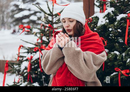 Eine junge Frau wärmt sich an einem kalten Wintertag die Hände, steht neben einem wunderschön geschmückten, mit Schnee bedeckten Weihnachtsbaum. Sie ist in einem weiß gekleidet Stockfoto