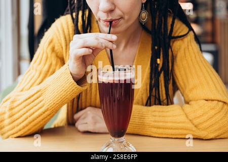 Eine Frau in einem gelben Pullover schlürft in einem gemütlichen Café ein erfrischendes Kaffeetränk durch einen Strohhalm. Der Fokus liegt dabei auf ihren Händen und dem Getränk Stockfoto