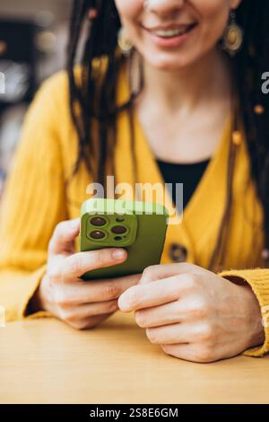 Eine Frau in einem senfgelben Pullover hält ein Smartphone mit einer grünen Hülle. Die gemütliche Atmosphäre und die Farben sorgen für eine warme und einladende Atmosphäre Stockfoto