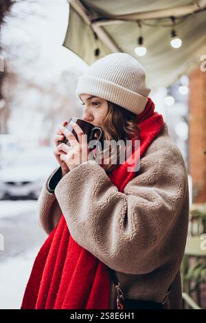 Eine junge, nachdenkliche Frau schlürft im Winter draußen ein warmes Getränk und trägt eine weiße Mütze und einen gemütlichen, braunen Mantel über einem knallroten Schal. Stockfoto