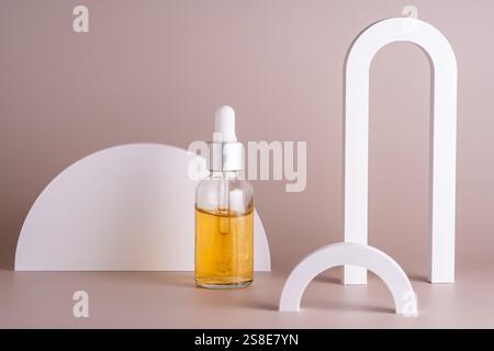 Eine Kosmetik in Glastropfflasche, gefüllt mit goldenem Öl, steht vor einem beigefarbenen Hintergrund, ergänzt durch weiße geometrische Formen, die eine Meile hervorheben Stockfoto