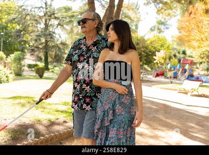 Ein blinder Vater mit einer Behinderung, der einen weißen Stock benutzt, wird von seiner fürsorglichen Tochter durch einen malerischen Park geführt. Zusammen verkörpern sie Liebe und Unterstützung. Stockfoto