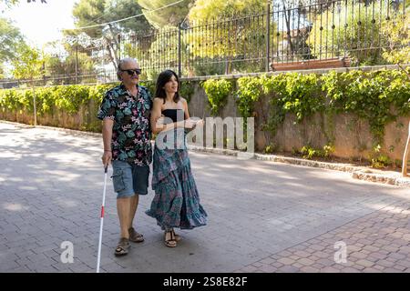 Fotografieren Sie einen blinden Mann mit einem weißen Stock, der von seiner unterstützenden Tochter an einem sonnigen Tag durch die Stadt geführt wird. Stockfoto
