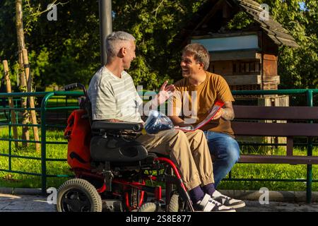 Zwei Freunde sitzen zusammen auf einer Parkbank, einer im Rollstuhl wegen Zerebralparese und der andere hält ein Notizbuch und führen ein lebhaftes Gespräch Stockfoto