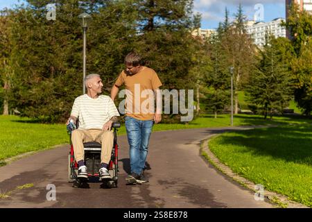 Ein junger Mann läuft neben einem älteren Rollstuhlfahrer mit Zerebralparese in einem Park und führt zu freundlichen Gesprächen. Der Weg ist umgeben von Gree Stockfoto