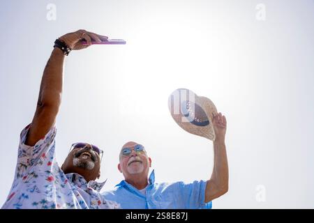 Von unten fängt ein fröhliches, schwules Paar ein Selfie ein, während es draußen steht. Beide Männer tragen eine Sonnenbrille und lächeln breit, was die fröhliche Stimmung verstärkt Stockfoto