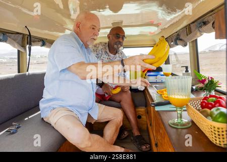 Ein älteres schwules Paar verbringt gute Zeit zusammen in einem Wohnmobil. Sie arbeiten mit der Zubereitung von Lebensmitteln und präsentieren einen gesunden Snack aus Bananen und Gemüse Stockfoto