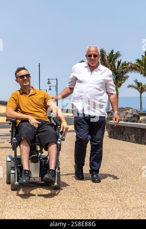Ein älterer Mann läuft mit einem erwachsenen Mann im Rollstuhl auf einer sonnigen Promenade. Beide Männer schauen zu und genießen einen gemütlichen Spaziergang zwischen Palmen Stockfoto