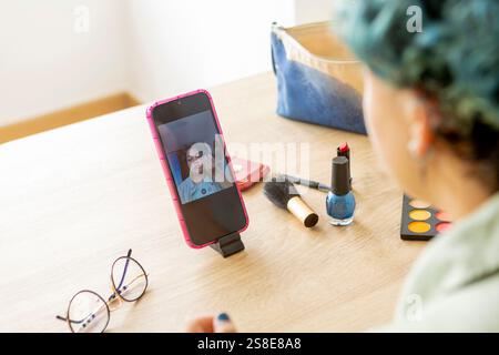 Ein Transgender nimmt mit seinem Handy ein Make-up-Tutorial auf und zeigt verschiedene kosmetische Produkte auf einem Tisch, darunter Pinsel, Lipsti Stockfoto
