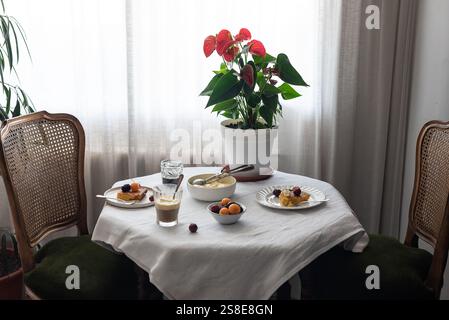 Warm beleuchteter Couchtisch mit einem weißen Tuch bedeckt, mit verschiedenen Desserts wie Kirschkuchen und Eis, einer Topfpflanze mit roter Blüte und einem Käsekuchen Stockfoto
