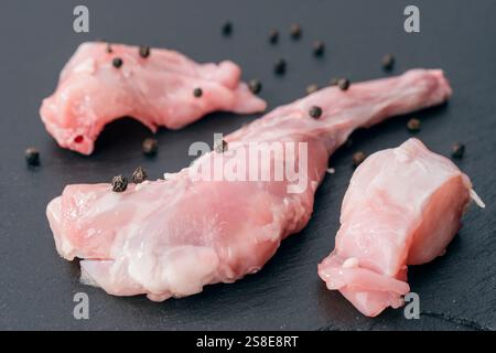 Nahaufnahme frischer roher Kaninchenfleischstücke auf dunklem Stein, bestreut mit ganzen schwarzen Pfefferkörnern, ideal für kulinarische oder Gourmet-Präsentationen und Speisen. Stockfoto