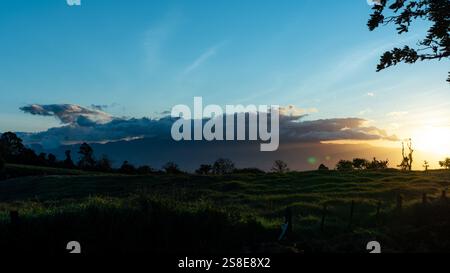 Erfassen Sie die ruhige Schönheit des Sonnenuntergangs über üppigen grünen Feldern unter einem lebendigen Himmel, der von der absteigenden Sonne beleuchtet wird, mit nebeligen Bergen in der Ferne. Stockfoto