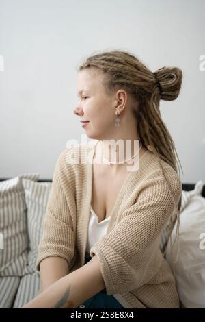 Eine junge Frau mit Dreadlocks und stilvollen Ohrringen sitzt auf einem bequemen Sofa. Sie trägt eine beige Strickjacke und ein weißes Oberteil und sorgt so für ein entspanntes und entspanntes Stockfoto