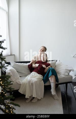 Mutter und Tochter teilen sich eine warme Umarmung auf einem gemütlichen gestreiften Sofa. Ein sanft beleuchteter Weihnachtsbaum in der Nähe der ruhigen Umgebung fängt den Geist von f ein Stockfoto