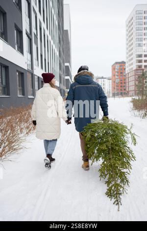 Ein Paar geht Hand in Hand durch ein schneebedecktes Stadtgebiet und trägt eine frisch geschnittene Weihnachtstanne. Sie sind warm gekleidet mit Mänteln und Hüten, Captu Stockfoto