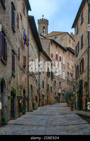 Eine charmante Kopfsteinpflasterstraße in Volterra, Toskana, gesäumt von traditionellen Steinhäusern mit Blumen. Mittelalterliche Architektur und Turmkrea Stockfoto