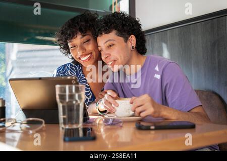 Ein fröhliches, multiethnisches LGBTQ+-Paar sitzt eng in einem gemütlichen Café und teilt einen warmen Moment bei einem Kaffee und einem Tablet. Sonnenlicht filtert durch das Fenster, c Stockfoto
