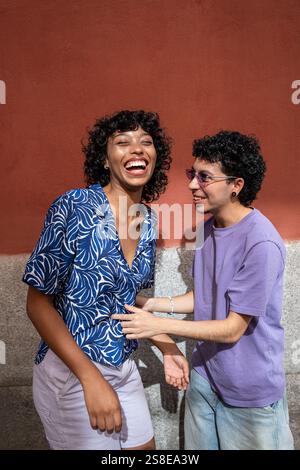 Ein multiethnisches LGBTQ+-Paar teilt einen freudigen Moment, während es draußen vor einer strukturierten, farbigen Wand lacht und Liebe und Vielfalt in feiert Stockfoto