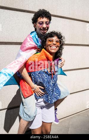 Ein lebhaftes, fröhliches, multiethnisches LGBTQ+-Paar genießt einen sonnigen Tag im Freien, umgeben von Stolz-Flaggen. Sie lächeln und umarmen, feiern Liebe und Stockfoto