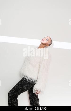 Eine Frau mittleren Alters mit rasiertem Kopf lehnt sich in einem stilvollen Ambiente selbstbewusst zurück. Sie trägt einen flauschigen weißen Mantel und eine schwarze Hose, die Eleganz verkörpert Stockfoto