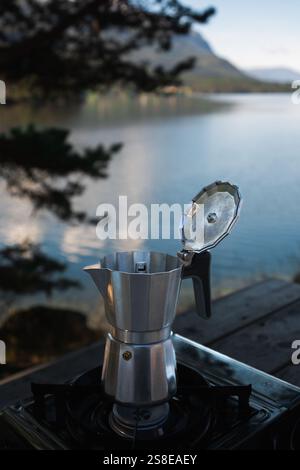Ein Mokakopf sitzt auf einem Campingofen neben einem ruhigen norwegischen See, umgeben von üppigen Bergen, perfekte Umgebung für ein entspannendes Outdoor-Abenteuer und Stockfoto