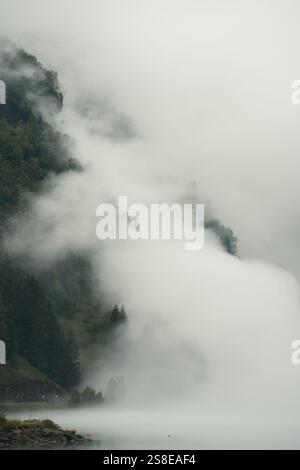 Dichter Nebel fließt über üppige, bewaldete Berge an einem ruhigen See in Norwegen. Die ätherische Szene fängt die raue Schönheit und ruhige Atmosphäre von Nordic lan ein Stockfoto