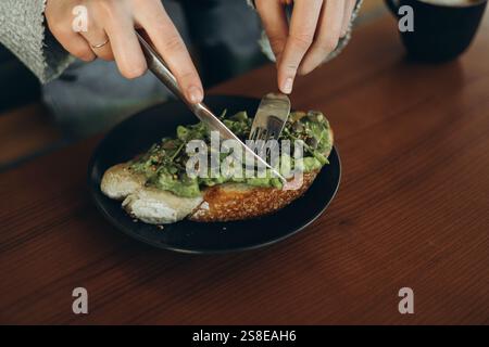 Nahaufnahme der Hände mit einer Gabel und einem Messer Avocadotoast auf einem dunklen Teller schneiden. Das offene Sandwich enthält frische, cremige Avocado auf einer Scheibe Stockfoto