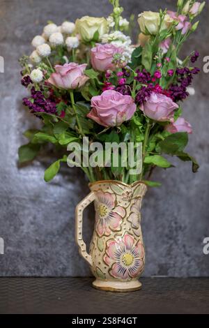 Wunderschöne Anordnung frischer rosafarbener, weißer und violetter Blumen in einer kunstvollen Keramikvase auf einer Arbeitsfläche mit neutraler strukturierter Rückseite Stockfoto
