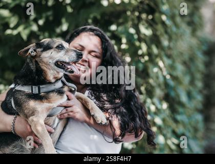 Latina-Mädchen hält einen mittelgroßen Hund in einem grauen Gurt. Sie hat lange dunkle Haare und trägt ein weißes T-Shirt. Hintergrund von grünem Laub im Freien Stockfoto