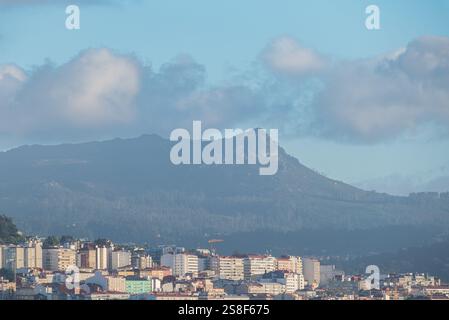 Vigos Gebäude vor einem Hintergrund von Bergen und einem leuchtend blauen Himmel mit verstreuten Wolken, die städtische und natürliche Schönheit zeigen Stockfoto