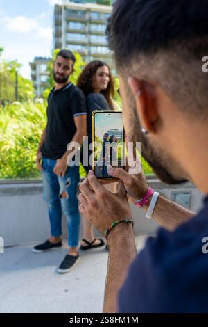 Ein junger Mann mit Bart fotografiert ein lächelndes Paar auf seinem Smartphone in einem Stadtpark. Das Paar steht im Hintergrund, rutschig Stockfoto