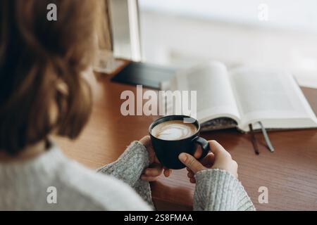 Eine entspannende Szene mit einer Person, die eine dampfende Tasse Kaffee hält, mit einem offenen Buch auf einem Holztisch, das eine friedliche Kaffeepause festlegt Stockfoto