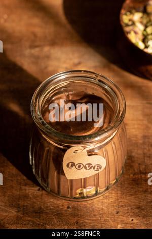 Ein warmer, sonnendurchfluteter Schuss veganer Schokoladenmousse in einem klaren Glas, verziert mit einem herzförmigen Liebesanhänger vor einem rustikalen Holzhintergrund. Stockfoto