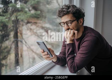 Erwachsener, gut aussehender Mann aus dem Nahen Osten mit Brille, scrollt morgens auf dem Handy und steht in der Nähe des Fensters. Stockfoto