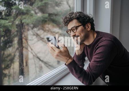 Junger gutaussehender Mann aus dem Nahen Osten mit Brille, der einen Videoanruf auf dem Handy hat, während er in der Nähe des Fensters steht. Stockfoto