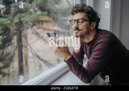 Junger gutaussehender Mann aus dem Nahen Osten mit Brille, der einen Videoanruf auf dem Handy hat, während er in der Nähe des Fensters steht. Stockfoto