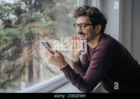 Junger gutaussehender Mann aus dem Nahen Osten mit Brille, der einen Videoanruf auf dem Handy hat, während er in der Nähe des Fensters steht. Stockfoto