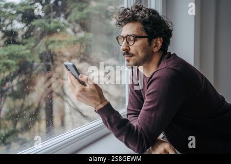 Erwachsener, gut aussehender Mann aus dem Nahen Osten mit Brille, scrollt morgens auf dem Handy und steht in der Nähe des Fensters. Stockfoto