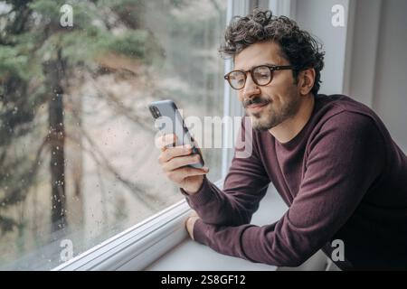 Erwachsener, gut aussehender Mann aus dem Nahen Osten mit Brille, scrollt morgens auf dem Handy und steht in der Nähe des Fensters. Stockfoto