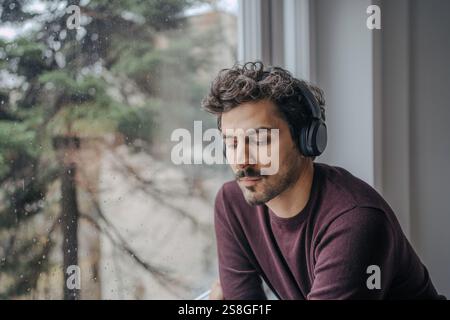 Gut aussehender Mann aus dem Nahen Osten mit On-Ear-Kopfhörern, Musik hören und sich in der Nähe eines Fensters entspannen. Stockfoto