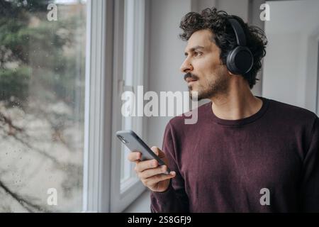 Gut aussehender Mann aus dem Nahen Osten mit On-Ear-Kopfhörern, Musik hören und sich in der Nähe eines Fensters entspannen. Stockfoto