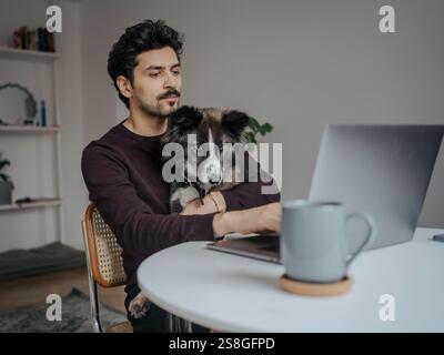 Junger, gutaussehender Mann aus dem Nahen Osten, der einen Computer benutzt und während der Arbeit einen süßen Hund im Schoß hält. Stockfoto