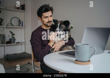Junger, gutaussehender Mann aus dem Nahen Osten, der einen Computer benutzt und während der Arbeit einen süßen Hund im Schoß hält. Stockfoto