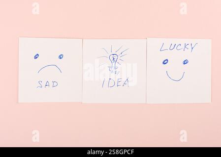 Drei Haftnotizen mit handgezeichneten Ausdrücken und Text: Traurig, Idee und Glück, skizziert in blauer Tinte auf einem sanften rosa Hintergrund Stockfoto