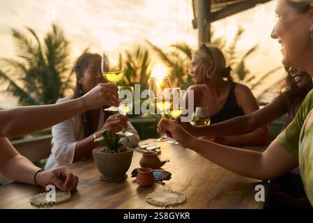 Verschiedene Frauen im tropischen Restaurant stoßen an. Freunde genießen Weinproben, fröhliche Stimmung und Blick auf den Sonnenuntergang. Geselliges Treffen, lebendiger Frauenclub. Feiern Stockfoto