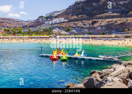 Malerischer Strand mit türkisfarbenem Wasser, farbenfrohen Tretbooten und Touristen, die sich unter Sonnenschirmen im sonnigen Küstenresort entspannen. Gran Canaria, Las Palmas, Spanien. Stockfoto