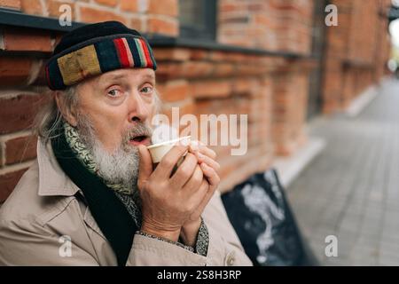 Nahaufnahme eines älteren Mannes, der draußen auf der Stadtstraße sitzt, warmes Getränk aus dem Pappbecher schlürft und Gefühle von Mitgefühl und Empathie weckt. Stockfoto